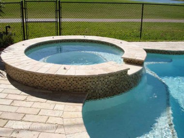 A raised circular spa has pale stone coping above mosaic sides, surrounded by rectangular pavers and a black garden fence.
