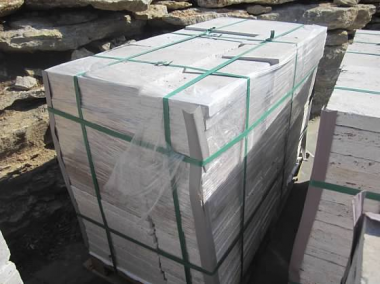 A pallet of pale stone pieces is wrapped in clear plastic and secured with green straps beside a rough stone wall.
